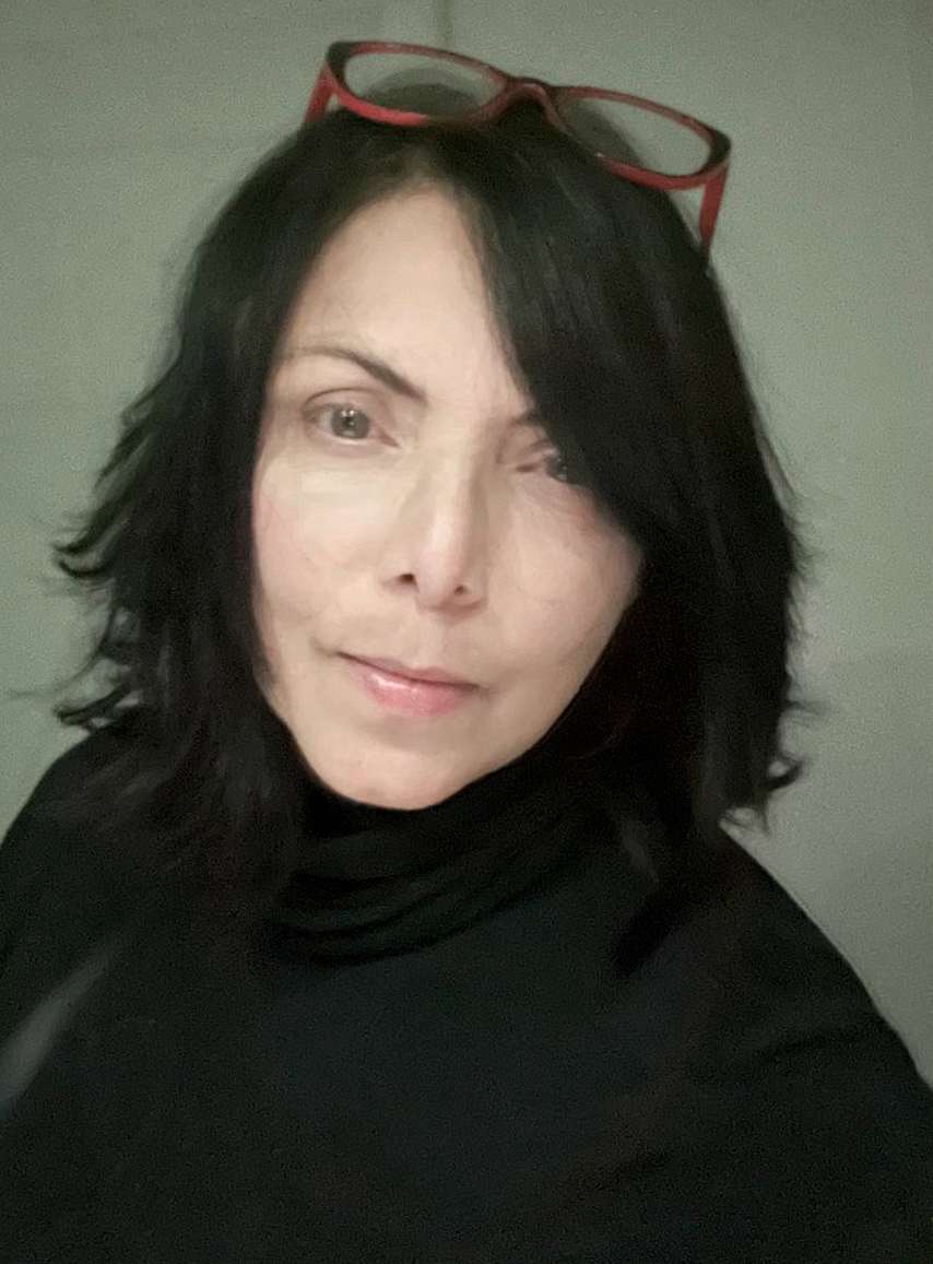 Simone Alves, sorridente, vestida de gola rolê preta, com rosto parcialmente coberto por longa franja preta, com óculos de leitura na cabeça.